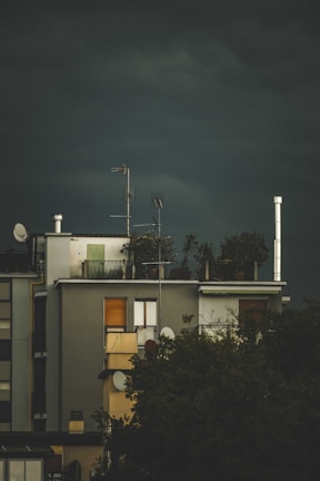 A building with a rooftop garden and several antennas. The structure appears modern, with large windows, and there are trees partially obscuring the lower part of the building. The sky is dark and overcast, suggesting an impending storm.