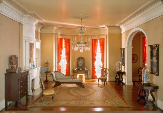 A classic Victorian-style room adorned with elegant antique furniture, including a chaise lounge, ornate chairs, and a detailed wooden table. The room features warm, striped wallpaper and two large windows with vibrant red curtains, allowing soft light to filter through. A grand chandelier hangs from the ceiling, adding a touch of sophistication. Several small decorative items are placed on the mantelpiece and small tables, contributing to the room's historical charm.