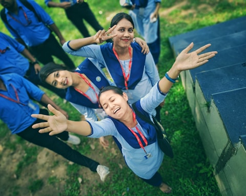 A group of young adults wearing blue uniforms with red lanyards are gathered outdoors. Three of them are prominently featured, posing playfully for the camera. The person in the front has arms outstretched and eyes closed, while the others are smiling and making peace signs. They stand on a grassy area near a set of steps.