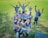 A group of young people wearing similar blue uniforms are standing together on a grassy field. They are displaying cheerful poses, with some raising their hands and others making peace signs. Each person is wearing a red lanyard with an identification card, and a few have backpacks.