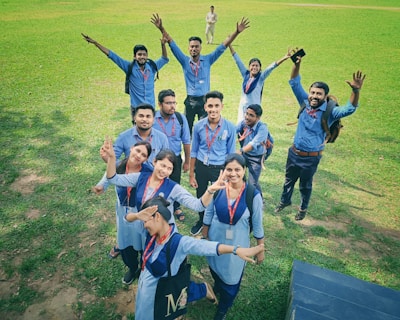 A group of young people wearing similar blue uniforms are standing together on a grassy field. They are displaying cheerful poses, with some raising their hands and others making peace signs. Each person is wearing a red lanyard with an identification card, and a few have backpacks.