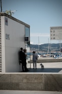 A waterfront scene with a marina in the background, featuring several boats docked. In the foreground, a person in light clothing is walking a small white dog. Another person in a suit is leaning against a building, talking to someone else. Signage is visible, indicating directions to various places.