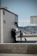 A waterfront scene with a marina in the background, featuring several boats docked. In the foreground, a person in light clothing is walking a small white dog. Another person in a suit is leaning against a building, talking to someone else. Signage is visible, indicating directions to various places.
