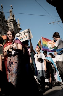 A group of people participating in a pride parade, holding a sign that says 'Born This Gay' with rainbow coloring on 'Gay.' Several participants are holding or wearing rainbow flags, and there is a historic building in the background.