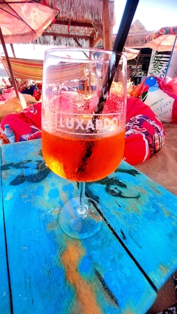 A vibrant scene of a beachside setting featuring a glass of orange-colored beverage on a blue wooden table. Surrounding this are colorful bean bags and sun umbrellas, along with a thatched roof structure casting shade.