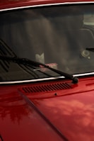 A close-up of a red car's front hood and windshield, featuring black wiper blades. The windshield reflects the sky and surrounding environment, while a pack of gum is visible on the dashboard. The car's surface is shiny, capturing subtle reflections.