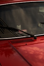 A close-up of a red car's front hood and windshield, featuring black wiper blades. The windshield reflects the sky and surrounding environment, while a pack of gum is visible on the dashboard. The car's surface is shiny, capturing subtle reflections.