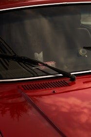 A close-up of a red car's front hood and windshield, featuring black wiper blades. The windshield reflects the sky and surrounding environment, while a pack of gum is visible on the dashboard. The car's surface is shiny, capturing subtle reflections.