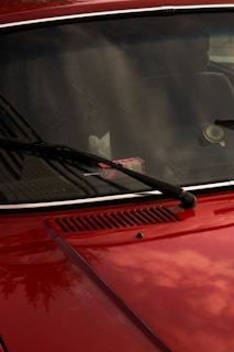 A close-up of a red car's front hood and windshield, featuring black wiper blades. The windshield reflects the sky and surrounding environment, while a pack of gum is visible on the dashboard. The car's surface is shiny, capturing subtle reflections.
