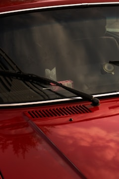 A close-up of a red car's front hood and windshield, featuring black wiper blades. The windshield reflects the sky and surrounding environment, while a pack of gum is visible on the dashboard. The car's surface is shiny, capturing subtle reflections.