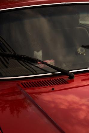 A close-up of a red car's front hood and windshield, featuring black wiper blades. The windshield reflects the sky and surrounding environment, while a pack of gum is visible on the dashboard. The car's surface is shiny, capturing subtle reflections.
