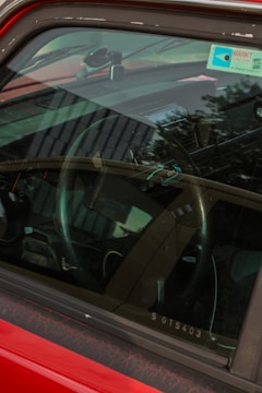 A close-up view through the window of a car's interior. The focus is on the steering wheel and dashboard, with reflections of trees and buildings visible on the window. A parking permit or similar sticker is seen on the windscreen.