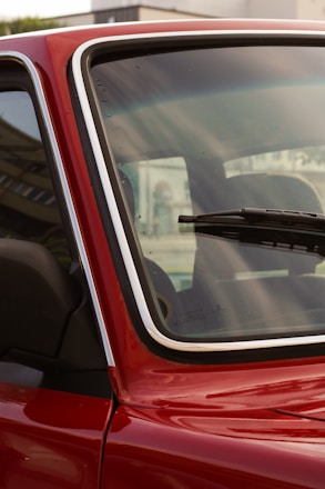 A close-up of a red car's windshield and part of the front door, highlighting the window and part of the interior. The emphasis is on the glass with droplets and the detail of the chrome trim. A windshield wiper is visible on the glass, as well as reflections and a blurred exterior backdrop.