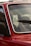 A close-up of a red car's windshield and part of the front door, highlighting the window and part of the interior. The emphasis is on the glass with droplets and the detail of the chrome trim. A windshield wiper is visible on the glass, as well as reflections and a blurred exterior backdrop.