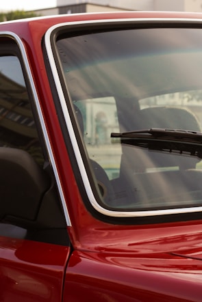 A close-up of a red car's windshield and part of the front door, highlighting the window and part of the interior. The emphasis is on the glass with droplets and the detail of the chrome trim. A windshield wiper is visible on the glass, as well as reflections and a blurred exterior backdrop.