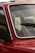 A close-up of a red car's windshield and part of the front door, highlighting the window and part of the interior. The emphasis is on the glass with droplets and the detail of the chrome trim. A windshield wiper is visible on the glass, as well as reflections and a blurred exterior backdrop.