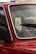 A close-up of a red car's windshield and part of the front door, highlighting the window and part of the interior. The emphasis is on the glass with droplets and the detail of the chrome trim. A windshield wiper is visible on the glass, as well as reflections and a blurred exterior backdrop.