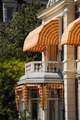 A building facade with several orange and white striped awnings over the windows and balcony. The structure features classic architectural elements with decorative balustrades and columns. Lush greenery surrounds the building, providing a picturesque backdrop.