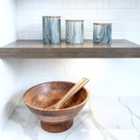 A modern kitchen setting with a wooden bowl and two utensils placed on a white marble countertop. Above the bowl, there is a wooden shelf holding three cylindrical canisters with a marbled gray design and wooden lids. The background features white subway tile backsplash.