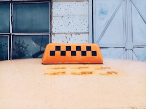 On top of a yellow vehicle, an orange taxi sign with a black checkered pattern is visible in the foreground. In the background, there are large glass windows and a weathered metal door.