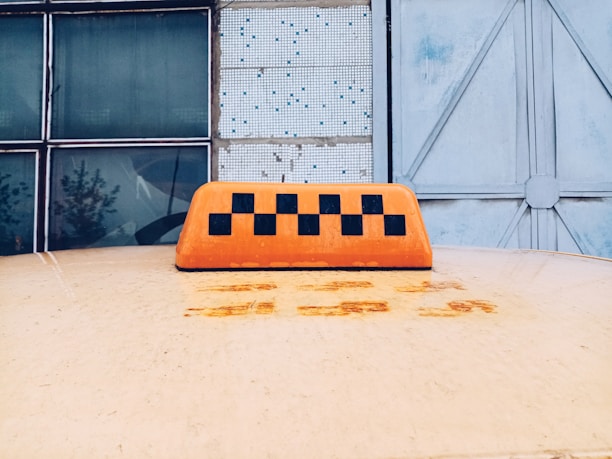 On top of a yellow vehicle, an orange taxi sign with a black checkered pattern is visible in the foreground. In the background, there are large glass windows and a weathered metal door.