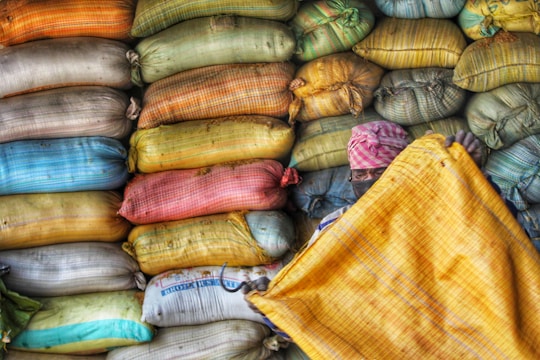 A person is partially visible holding a large yellow sack or sheet in front of a wall of stacked, multi-colored bags. The bags are arranged neatly in rows, creating a vibrant and textured background. The individual appears to be wearing a pink patterned headscarf and a face covering.