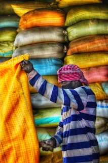 A person wearing a striped shirt and a pink and white checkered headscarf is handling a large yellow sack. Behind them, there are stacks of colorful bags in various shades of yellow, orange, blue, and gray.