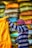 A person wearing a striped shirt and a pink and white checkered headscarf is handling a large yellow sack. Behind them, there are stacks of colorful bags in various shades of yellow, orange, blue, and gray.