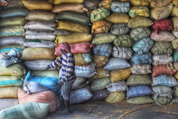 A person wearing a striped shirt and headscarf is standing next to a large stack of colorful, multi-sized bags filled with what appears to be grain or produce. The bags are stacked neatly against a wall, and some grain seems to be scattered on the ground.