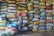 A person wearing a striped shirt and headscarf is standing next to a large stack of colorful, multi-sized bags filled with what appears to be grain or produce. The bags are stacked neatly against a wall, and some grain seems to be scattered on the ground.