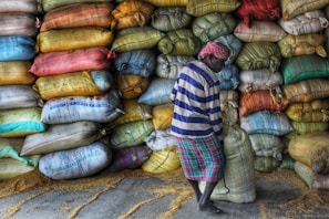 A person wearing a striped shirt and a headscarf is lifting a sack in front of a large stack of colorful sacks. The sacks are piled high against a wall and appear to contain grains, as some grains are scattered on the floor.