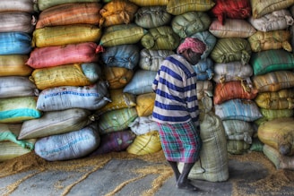 A person wearing a striped shirt and a headscarf is lifting a sack in front of a large stack of colorful sacks. The sacks are piled high against a wall and appear to contain grains, as some grains are scattered on the floor.
