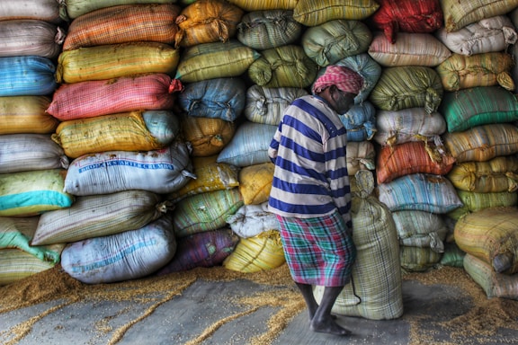 A person wearing a striped shirt and a headscarf is lifting a sack in front of a large stack of colorful sacks. The sacks are piled high against a wall and appear to contain grains, as some grains are scattered on the floor.
