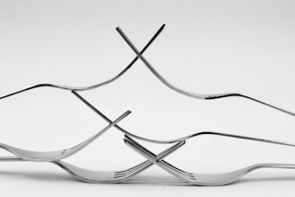 An artistic arrangement of several intertwined and balanced forks creates an intricate geometric pattern. The forks appear to be made of metal, displaying a shiny, reflective surface.