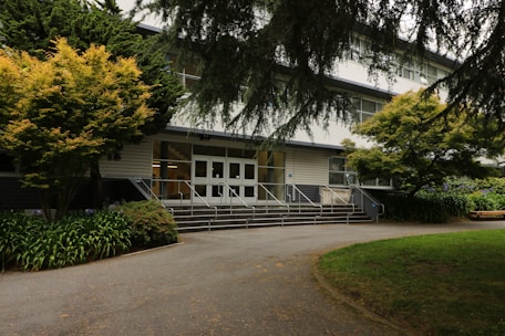 A multi-story building entrance flanked by trees and greenery. The path leading up to the entrance is bordered by well-maintained plants. The white facade of the building is contrasted with darker window frames and the area is shaded by tree branches.