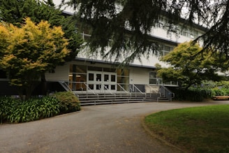 A multi-story building entrance flanked by trees and greenery. The path leading up to the entrance is bordered by well-maintained plants. The white facade of the building is contrasted with darker window frames and the area is shaded by tree branches.