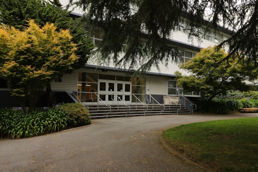 A multi-story building entrance flanked by trees and greenery. The path leading up to the entrance is bordered by well-maintained plants. The white facade of the building is contrasted with darker window frames and the area is shaded by tree branches.