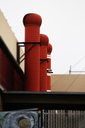Several large red industrial vents or pipes are attached to the side of a building. These cylindrical structures have ribbed textures and are positioned vertically along the wall. The background includes parts of the building and a clear sky.