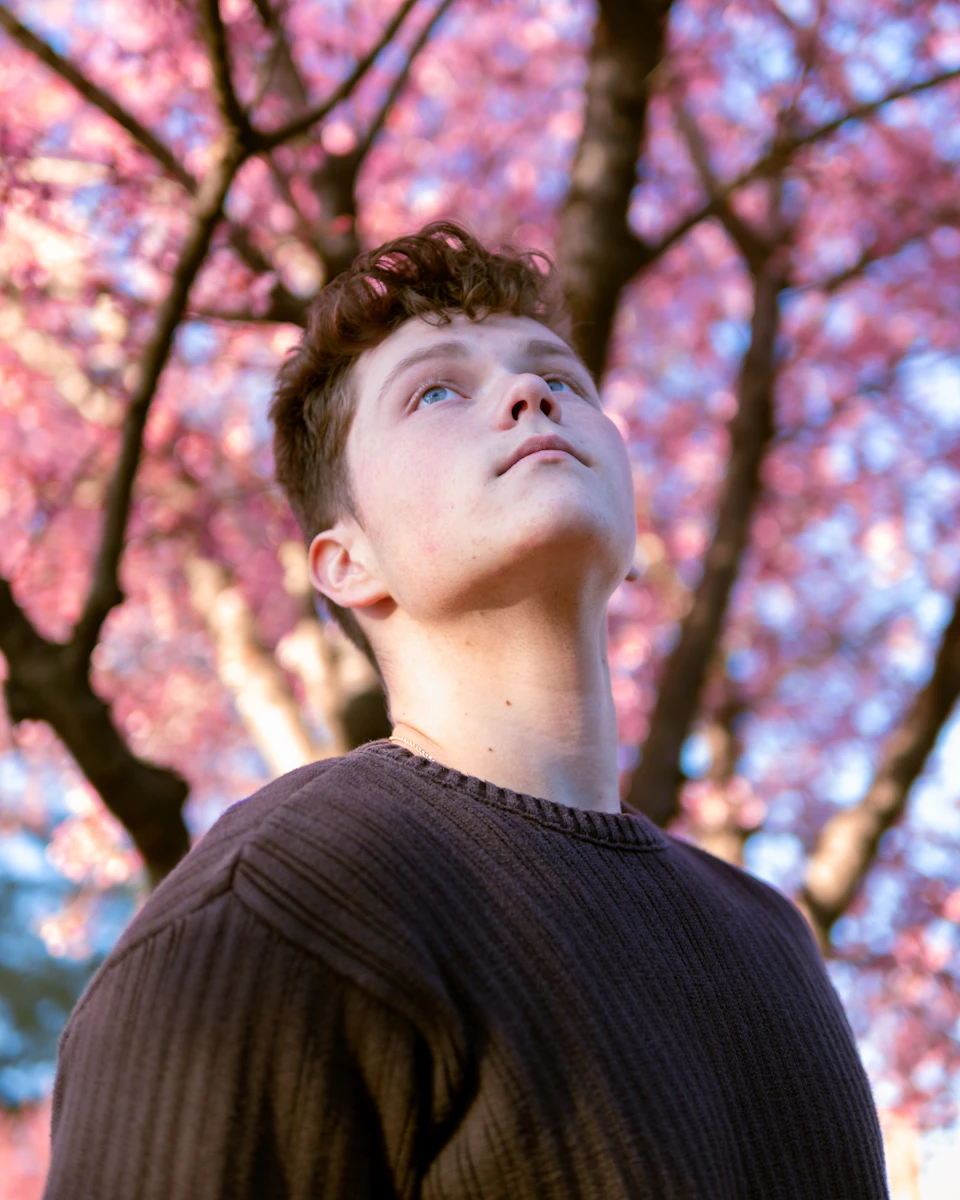Young man stands under tree with pink flowers, looking up at the sky outdoors