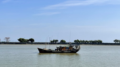 A small fishing boat with equipment and antennas is anchored on a calm body of water. The horizon shows a tree-lined embankment under a clear blue sky.