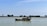 A small fishing boat with equipment and antennas is anchored on a calm body of water. The horizon shows a tree-lined embankment under a clear blue sky.