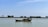 A small fishing boat with equipment and antennas is anchored on a calm body of water. The horizon shows a tree-lined embankment under a clear blue sky.