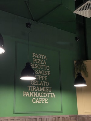 A green wall with a menu list featuring Italian food items such as pasta, pizza, risotto, lasagne, soups, gelato, tiramisu, panna cotta, and coffee. Three black pendant lights hang from the ceiling, casting a warm glow over the area. There is also a glimpse of some greenery in the form of palm leaves.