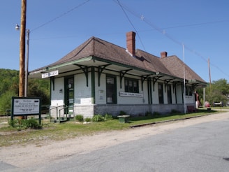A historic railway station with green and white wooden exterior, featuring a sloped roof and multiple windows. The building is labeled Gorham and Grand Trunk Railway. A sign nearby indicates it is part of the Gorham Historical Society and Railroad Museum. The surroundings include a gravel path, some greenery, and utility poles.