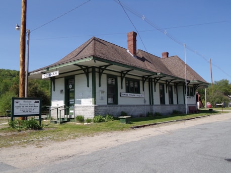 A historic railway station with green and white wooden exterior, featuring a sloped roof and multiple windows. The building is labeled Gorham and Grand Trunk Railway. A sign nearby indicates it is part of the Gorham Historical Society and Railroad Museum. The surroundings include a gravel path, some greenery, and utility poles.