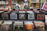 A collection of vinyl records is displayed in black crates on shelves. The surroundings include various music paraphernalia such as CDs, posters, and decorative items. There are also some framed artworks and magazine covers on display, contributing to a vintage and eclectic atmosphere.