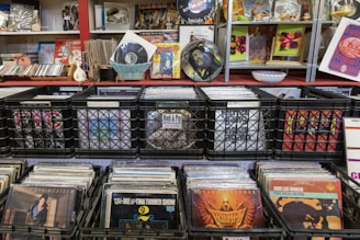 A collection of vinyl records is displayed in black crates on shelves. The surroundings include various music paraphernalia such as CDs, posters, and decorative items. There are also some framed artworks and magazine covers on display, contributing to a vintage and eclectic atmosphere.