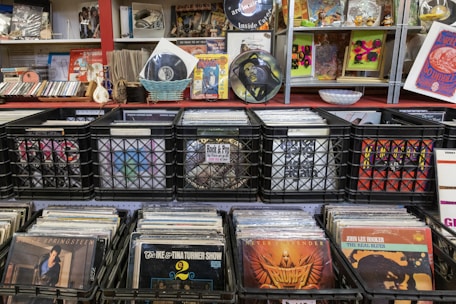 A collection of vinyl records is displayed in black crates on shelves. The surroundings include various music paraphernalia such as CDs, posters, and decorative items. There are also some framed artworks and magazine covers on display, contributing to a vintage and eclectic atmosphere.