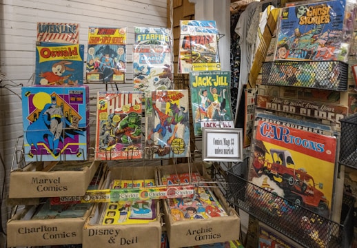 A collection of vintage comic books and magazines is displayed on metal racks. Titles like Oswald, Spider-Man, Hulk, and Jack and Jill are visible. The comics are enclosed in plastic sleeves, and some racks are labeled with categories like Marvel Comics, Charlton Comics, Disney & Dell, and Archie Comics. A sign indicates that the comics/mags are priced at $3 unless marked otherwise. There are various colors and illustrations on the covers, showcasing different characters and themes.