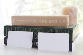 A small stack of books is placed on a light-colored surface near a window. The top book has a brown cover with the title 'The End' and is part of 'A Series of Unfortunate Events.' Below it is a dark green book with a decorative cover. Two blank white index cards are leaning against the books. In the background, a light brown candle is visible.